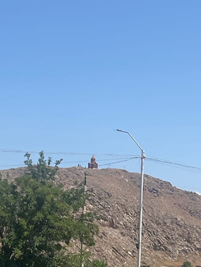 The image depicts a mountainous landscape with a clear blue sky. A rocky cliff face dominates the right side of the frame, with sparse green foliage at its base. A metal pole, possibly part of a utility structure or lighting fixture, extends horizontally across the middle ground. Thin wires are strung tautly above and in front of the cliff. In the distance, perched on the upper part of the cliff, a small building with a distinctive brown, conical roof is visible. The scene appears to be set in a natural environment, likely during the daytime given the bright sky. The location is identified as Surenavan, Armenia, suggesting a terrestrial, potentially arid or semi-arid, mountainous region. No people or specific activities are evident. The overall impression is one of a serene, somewhat rugged natural setting with man-made infrastructure present.
