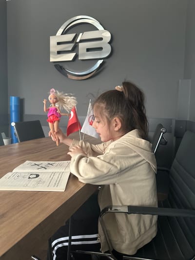 A young female child is seated at a large, rectangular wooden table within an office or meeting room located in Kemalpaşa, Turkey. The child, with light brown hair tied in a high ponytail and wearing a beige hooded jacket over dark pants with white stripes, holds a blonde doll, dressed in a pink top and red skirt, elevated with her left hand. The doll's right arm is raised. An open book featuring black and white illustrations and text, including prominent "X X" markings, rests on the table in front of the child. To the right of the doll, a small Turkish flag and another white flag with a red symbol are displayed on the table. The child's gaze is directed towards the doll. In the background, a large, metallic, reflective logo displaying the letters "EB" within an oval is mounted on a dark grey wall. Black office chairs with metal frames are visible around the table, and a partial view of a light blue cylindrical object is present on the far left.