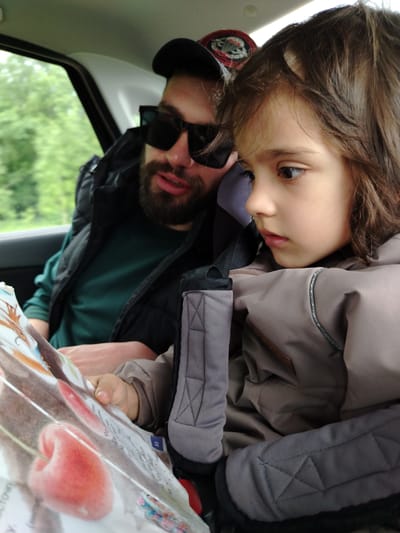 A photograph shows a man and a young child in a car. The man is wearing a black cap, black sunglasses, a green shirt, and a black vest. He has a beard. The child is seated in a car seat and is wearing a beige jacket. Both the man and the child are looking at a book with a picture of a cherry on it. The background shows greenery outside the car window. The location is Краснояр, Russia.