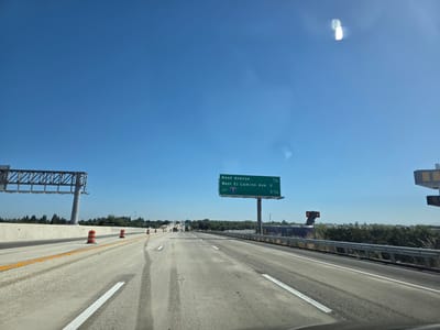 This media file captures a view from a vehicle driving on a highway in West Sacramento, USA. The scene is set under a clear, bright blue sky, suggesting a sunny day with good visibility, likely midday.
The primary focus is the highway itself, with multiple lanes visible. The road surface appears to be concrete, showing some tire marks and wear. Orange construction barrels are placed along the shoulder, indicating potential roadwork or lane closures. A concrete barrier separates the lanes from the opposing traffic. Overhead, a large highway sign structure is visible on the left, and further ahead, another prominent green highway sign provides directional information.
The sign reads: "Reed Avenue 1/4", "West El Camino Ave 2", and "JCT 5 3 1/4". This text indicates upcoming exits and distances, with "JCT 5" suggesting a junction with Interstate 5.
In the distance, trees and vegetation line the sides of the highway, with some industrial or commercial buildings also visible. A few other vehicles are seen on the road ahead, moving in the same direction. The overall impression is a typical highway scene during daylight hours, with clear weather and no unusual activity. The presence of construction barrels suggests an ongoing infrastructure project.