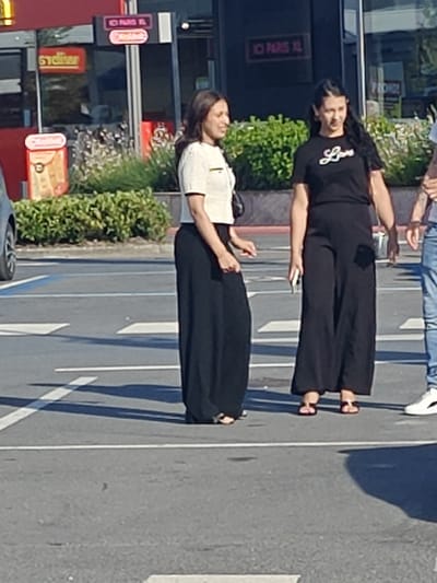 The image shows three people standing on a paved area with white lines, possibly a parking lot or a pedestrian crossing. The setting appears to be outdoors, with some greenery and buildings in the background.
On the left, a woman is wearing a white, short-sleeved top and a long, flowing black skirt. She is looking towards her right and has her left hand slightly raised.
In the center, another woman is wearing a black t-shirt with the word "Love" written in white script across the chest, and a long, wide-legged black skirt. She is holding a white object, possibly a phone, in her right hand and is looking forward.
On the right, a person in blue jeans and white sneakers is partially visible. They are standing and looking towards the left.
In the background, there are buildings with signage. One sign in red letters reads "STADION". Another sign above it reads "ICI PARIS XL". The lighting suggests it is daytime, likely sunny, given the shadows. The overall mood appears casual. The location context provided is Sint-Pieters-Leeuw, Belgium.