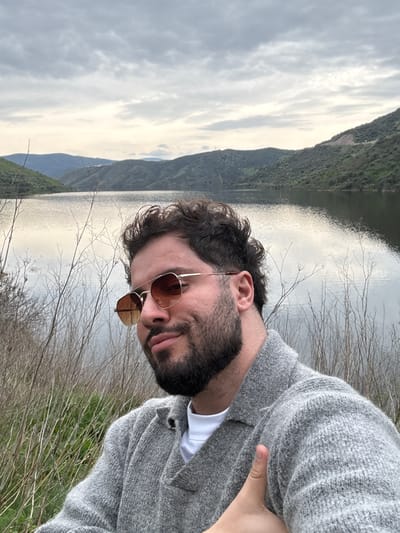 A male individual is depicted in a medium close-up selfie in a natural outdoor environment near Mogadouro, Portugal. He has dark, curly hair, a full beard, and is wearing gold-framed aviator-style sunglasses with brown-tinted lenses. He is dressed in a textured light gray knitted cardigan over a white crew-neck t-shirt. His right hand is raised, performing a "thumbs-up" gesture, visible against his sweater in the lower right portion of the frame. He has a slight smile and is looking towards the camera. The background features a large, calm body of water, likely a reservoir or river, which reflects the overcast sky. Beyond the water, vegetated hills and mountains rise, receding into the distance under a uniformly cloudy, light gray sky. The immediate foreground includes dry, thin, brown plant stalks and some underlying green foliage.