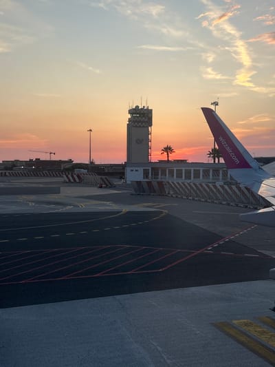 The image shows an airport tarmac at sunset. A Wizz Air airplane wing with "wizzair.com" written on it is visible in the upper left quadrant. In the background, a control tower stands against a sky filled with soft orange and pink hues, indicating the time of day is sunset. Two palm trees are visible near the control tower. The tarmac is marked with yellow and red lines, typical of airport runways and taxiways. In the middle ground, there is a barricade with red and white stripes. A crane can be seen in the far background. The overall scene evokes a sense of calm and transition associated with the end of the day. The location is identified as Lamezia Terme, Italy.