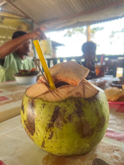The image displays a green coconut, serving as a beverage, centrally positioned on a patterned table. The top of the coconut is cut open, revealing the inner white husk, with several husk fragments remaining around the opening. A yellow plastic straw and a silver-colored spoon are inserted into the coconut. The coconut's exterior is mottled green and brown, and small droplets of liquid are visible on its surface and on the table. In the blurred background to the left, an out-of-focus male individual with dark skin, wearing a light green shirt, is seated, with his left arm raised. A dark bowl and a white bowl containing food are present on the table in front of him. To the right, another heavily out-of-focus individual is seen from behind, next to a glass of dark liquid containing a red straw, and a plate holding light-colored fried food items. The environment is an open-air or semi-enclosed dining area with a corrugated metal roof overhead, illuminated by bright natural light, situated in Gembong, Indonesia.