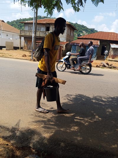 In Egbedi, Nigeria, a man walks on the right side of a paved road, carrying a black bucket and a piece of wood. He is wearing a yellow t-shirt and dark shorts, with a backpack slung over his left shoulder. His feet are bare except for one sandal on his left foot. In the background, a motorcycle with two passengers travels away from the viewer on the same road. The environment is urban, with buildings featuring corrugated metal roofs visible on the left and right sides of the street. Trees with dappled sunlight cast shadows on the road. The sky is partly cloudy.