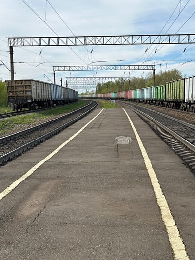 The image depicts a railway platform in Кечёво, Russia. Two sets of train tracks curve through the frame. On the left of the platform, a train with multiple cargo cars is parked, while on the right, a solid white line marks the edge of the platform. Overhead, power lines are suspended by a series of metal towers. The platform itself is paved with asphalt.