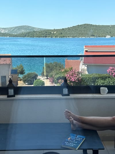 The image depicts a serene balcony overlooking the Adriatic Sea in Grad Šibenik, Croatia. A person's legs and bare feet are visible, crossed and resting on a dark table. On the table lies a book titled "I'm Laughing Because I'm Crying" by Youngmi Mayer. In the foreground, on the balcony ledge, are two black lanterns with white candles inside and a white mug. Beyond the balcony, the crystal-clear blue water stretches to a green, tree-covered island, with a few sailboats dotting the horizon. Two houses with red tile roofs are partially visible on the left and right sides of the scene, separated by lush green foliage and pink flowering bushes. The sky is a clear, bright blue, indicating a sunny day.