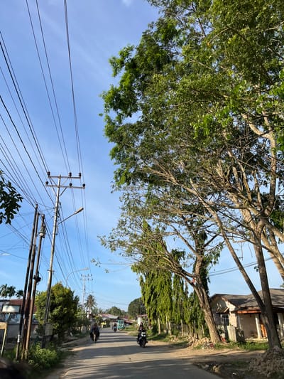 The image depicts a street scene in Alurcucur, Indonesia, under a clear blue sky. Power lines stretch across the frame, supported by utility poles. On the left side of the road, there are several poles, some with what appear to be electrical transformers and streetlights. Adjacent to these poles is a building with weathered walls and a dark roof, adorned with signage. Further down the street, more utility poles and trees line the road.
Several individuals are on motorcycles, traveling along the paved road. One motorcycle, ridden by a person wearing a blue shirt, is in the foreground on the left. Another motorcycle, with a rider in a dark shirt, is further ahead on the right. In the distance, a light blue van or bus is visible. The environment suggests a rural or suburban setting with residential buildings and abundant vegetation, including tall trees with dense foliage. The lighting indicates it is daytime, with sunlight casting shadows on the road.