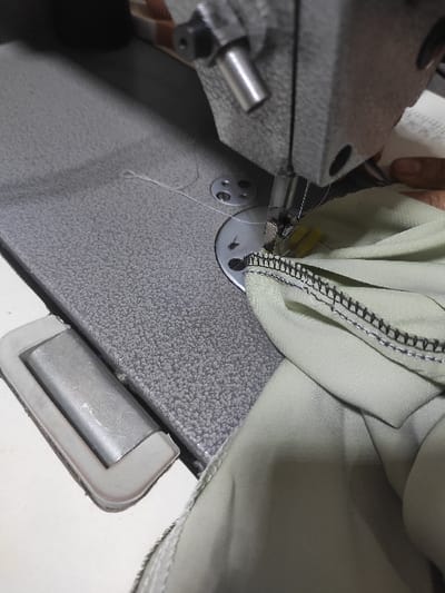 The image depicts a close-up, overhead view of an industrial sewing machine in operation, located in Sidi Chahmi, Algeria. A human hand, characterized by light brown skin, is visible on the right side of the frame, guiding a piece of light green fabric under the machine's presser foot. The sewing machine features a grey, textured metal bed, a prominent needle mechanism, and a white thread actively engaged in the stitching process. The light green fabric, likely a garment, is being finished along its edge with a distinct black serger stitch, forming an overlock seam. A metal component encased in a white rubber frame, potentially part of a foot pedal or an accessory, is situated on the lower left of the machine's bed.