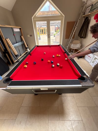 A man is playing pool in a room in a house located at 54.025 latitude and -0.933 longitude in Sand Hutton, United Kingdom. He is standing near a red pool table with a wooden cue stick, preparing to hit a ball. There are several billiard balls scattered across the red felt table. The room has a tiled floor. A large window with French doors is visible in the background, showing a view of a garden. On the right side of the room, a ladder and a small white chair with a cushion are visible. A dark, rectangular object, possibly a table tennis table, is also visible on the left side of the image. There are several items on the shelves on the wall behind the man. The house is in a residential area.