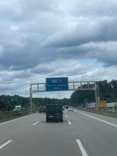 The image shows a highway scene on a cloudy day. A blue overhead traffic sign indicates "Dreieck Spreau 1800 m" with an arrow pointing to the right and a highway number 8 within a circle. The highway is a multi-lane road with cars and trucks driving on it. The road is lined with trees and vegetation on both sides. In the foreground, a black van with German license plates is visible. In the background, several trucks and trailers are parked along the side of the road. The overall scene suggests an approach to a highway junction or interchange. The location is identified as Königs Wusterhausen, Germany.