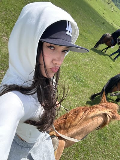 A medium-angle, eye-level outdoor shot shows a young woman wearing a white hoodie and a black New York Yankees baseball cap, positioned at the bottom left. She is angled towards the right. Her dark hair flows across the image, partially obscuring the view. She is riding a light brown horse, visible from the neck up, with a saddle and reins. Two other people are seen riding horses in the background, with one horse being dark brown and the other obscured. They are on a green grassy field. The location is Teghut, Armenia.