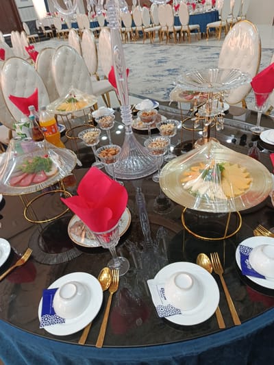 This media file captures a formal dining setup, likely for a wedding reception or similar celebratory event. The scene is set in what appears to be a banquet hall, with a long, elegantly decorated table as the focal point. The table is laden with multiple place settings, each featuring white plates, golden cutlery (forks and spoons), and small white bowls. Red napkins are prominently displayed at each setting, folded in a distinctive tulip shape.

Several tiers of crystal and gold serving stands are arranged on the table, holding various food items. One stand displays sliced meats and greens, covered with plastic wrap. Another holds a selection of cheeses and what appear to be fruit slices, also under plastic. Smaller glass cups contain nuts, suggesting appetizers or snacks. A tall, ornate crystal centerpiece dominates the background.

The chairs surrounding the table are plush and white with golden accents, indicating a luxurious setting. In the background, more tables are visible, set with blue tablecloths and matching chairs, suggesting a large gathering. The lighting appears to be artificial, casting a warm glow, and the overall atmosphere conveys a sense of elegance and celebration. The visible text on a small white packet reads "ELLUM". The location context is Shahrisabz City, Uzbekistan.