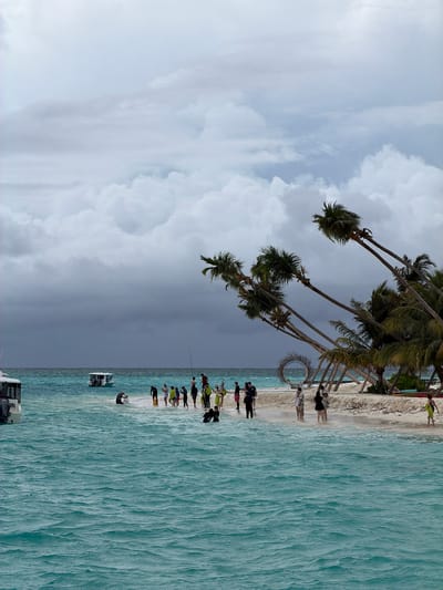 The image depicts a beach scene in Fulidhoo, Maldives. Palm trees are inclined towards the ocean. A group of people is standing on the white sandy beach, some near a circular photo booth. The turquoise water extends into the distance. Two boats are visible near the shore. The sky is cloudy with varying shades of gray.