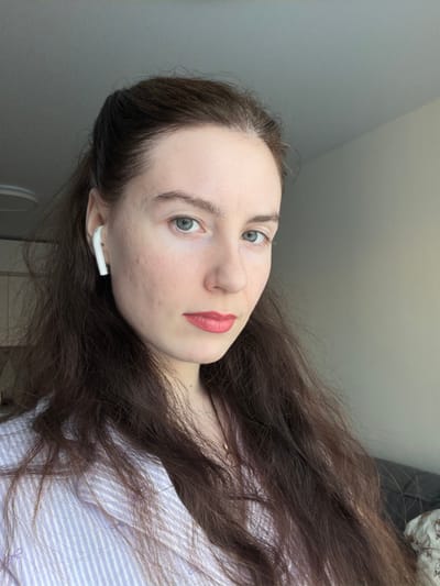 The image displays a head-and-shoulders portrait of an adult female located indoors in what is identified as the city of Ковалёво, Russia. The individual has a light complexion, dark brown wavy hair extending past her shoulders, and blue-grey eyes. She is wearing red lipstick. Visible acne scarring is present on her right cheek. A single white wireless earbud is inserted into her right ear. She is dressed in a light blue and white striped button-up shirt, and a thin gold-colored necklace with a small pendant is visible. Her gaze is directed towards the camera. The background consists of a plain light grey ceiling and a light beige wall. An indistinct dark object is partially visible in the lower right background, and a circular ceiling fixture is partially visible in the upper left background.