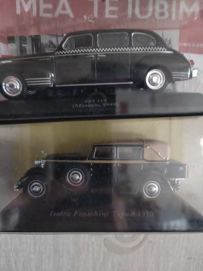 The image displays two model cars, one above the other. The upper model is a black ZIS 110 taxi from Moscow, 1948. It features a checkered band along the roofline and chrome detailing. The lower model is a black and tan Isotta Fraschini Tipo 8 from 1930, with a convertible top. Both models are presented in plastic packaging. The background shows a red sign with white lettering that reads "MEA TE TUBIM", and to the right, part of a website address is visible, ending with "sters.com". The scene appears to be an indoor display, possibly a shop or a collection. There is no indication of the city of Soisy-sous-Montmorency, France, or any specific location within it. The image does not contain any people.