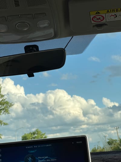 The image shows the interior of a car looking out through the windshield at a bright blue sky filled with fluffy white clouds. The car's rearview mirror is visible, as is the car's overhead console with various buttons and lights. A sun visor with safety warnings, including an airbag symbol and a graphic indicating no children in the front seat, is also present. The car's dashboard display screen shows the time as 18:29 and song information, including the title "Revenge" by Belchev, Billy Blake. Temperature readings of 22.0°C inside and 26.5°C outside are also displayed. The location is identified as Novomar'evskaya, Russia. Trees and a street light are visible in the background outside the car.