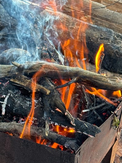 This is a close-up, eye-level shot of a bonfire in a rusty metal brazier, likely located in Fort-Krasnaya Gorka, Russia. The fire is actively burning, with orange and yellow flames licking at dark, charred logs and smaller twigs. Wisps of pale blue smoke curl upwards from the left side of the frame, partially obscuring some of the burning wood. The brazier itself is made of weathered metal, with visible rust and a slightly angled opening at the bottom. The background is out of focus, showing what appears to be more logs stacked horizontally. There are no people or animals visible in the image.