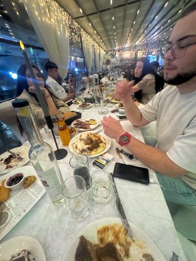 The image shows a long table at a restaurant, possibly outdoors under a retractable awning illuminated with string lights. The table is covered with a white tablecloth, and numerous people are seated along it. Food and drinks are scattered across the table, including partially eaten plates of food, empty wine bottles, glasses, and cutlery. A person wearing a white t-shirt and a red watch is prominently positioned in the foreground. Another person, wearing glasses, is positioned near the top of the image. The general setting suggests a social gathering or meal at a restaurant. I can not determine its location in Zlati Voyvoda, Bulgaria.