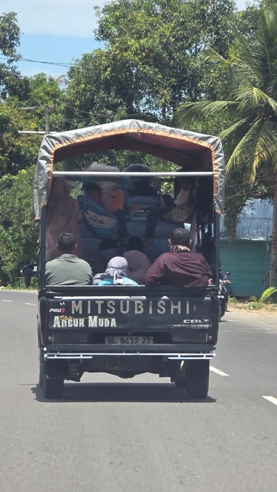 The image depicts the rear of a Mitsubishi Colt L300 pickup truck in Gampongrayadagang, Indonesia. The vehicle is black and filled with people, sitting in the open truck bed. The truck has a frame structure covered with a canvas or tarp material overhead. The lettering "MITSUBISHI" and "Ancuk Muda" are visible on the back of the truck. The license plate reads "BL 8412 ZT". The vehicle is driving on an asphalt road, with trees and vegetation visible on either side. Some buildings are visible in the background.