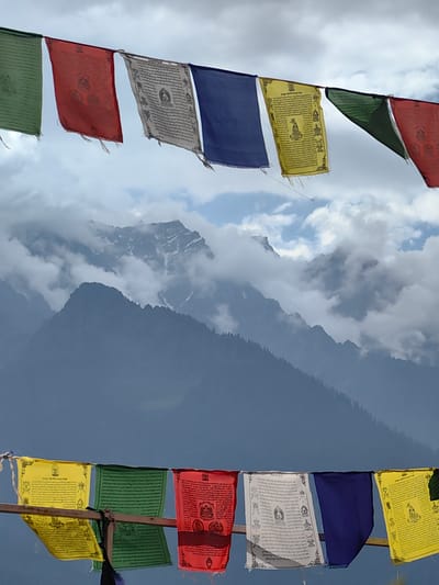 In Siumsa, India, prayer flags are strung across a vista of misty mountains under a cloudy sky. The flags, featuring Tibetan script and imagery, are a colorful addition to the dramatic landscape. The upper row of flags includes green, red, white, blue, and yellow, while the lower row displays yellow, green, red, white, and blue. The mountains are layered in the background, with some peaks partially obscured by clouds, and trees dot the slopes of the closer mountains.