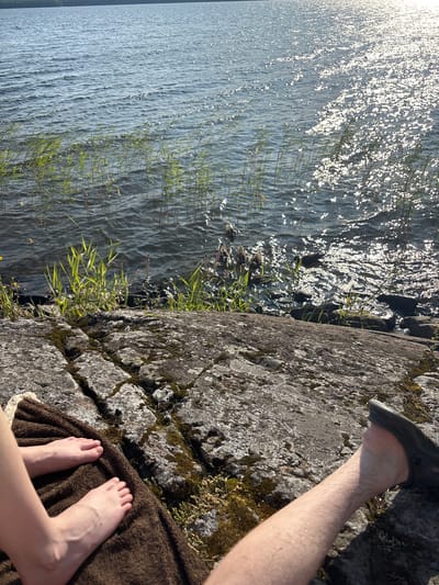 In the city of Shuiskaya Chupa, Russia, a close-up shot captures two individuals relaxing on a rocky shore by a body of water. The foreground is dominated by the texture of the grey, moss-covered rocks and a brown, plush towel. On the towel, a pair of bare feet, belonging to one individual, are visible, with pale skin and neatly trimmed toenails. To the right, another individual's leg, clad in a dark, perforated shoe, rests on the rocks, revealing slightly hairy skin. The water, reflecting the bright sunlight, shimmers with a multitude of sparkling highlights. Reeds and grasses grow along the water's edge, and a group of ducks can be seen swimming in the distance. The overall scene depicts a tranquil outdoor setting, likely a summer day by the lake.