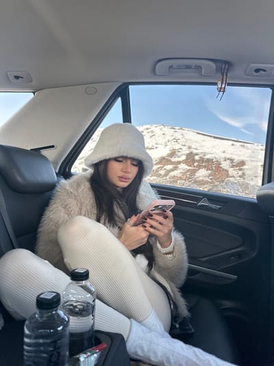 A female individual is seated in the rear passenger seat of a vehicle, in what is identified as the city of Jraber, Armenia. She is wearing a white faux fur bucket hat, a light-colored shaggy faux fur coat, and ribbed off-white leggings. Her dark hair is visible beneath the hat, and her fingernails are painted dark red. She is holding a light pink-cased smartphone in both hands and looking down at its screen. Two clear plastic water bottles are visible in a cup holder in the foreground. Sunglasses are hanging from a passenger grab handle above the right rear window. Through the vehicle's windows, a snow-covered, rocky landscape under a clear blue sky is visible, indicating a cold, bright environment.