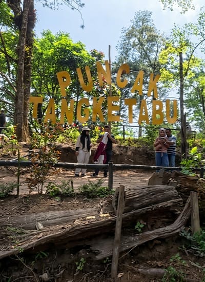 This image captures an outdoor scene in Laugumba, Indonesia, featuring a large yellow sign with the text "PUNCAK TANGKETABU" prominently displayed. The sign is constructed of individual letter shapes mounted on poles, set against a backdrop of lush green trees and a bright blue sky. In the foreground, a dirt path is visible, bordered by a wooden railing. Several people are present in the mid-ground, some standing near the sign and others walking along the path. One woman, wearing a light-colored hijab and a long tunic, stands near the sign. A man beside her is holding a red and white striped cloth. Further to the right, a couple is interacting; the woman, in a pink hijab, is looking at her phone, while the man, wearing a striped shirt, stands next to her. The environment appears to be a natural, wooded area, possibly a park or recreational spot, with fallen logs and vegetation scattered around the path.