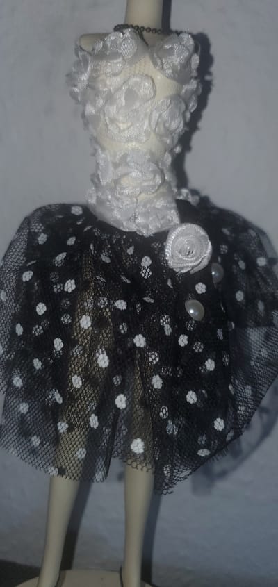 A close-up, high-angle, slightly blurry photograph shows a mannequin with a doll-sized black polka dot skirt and a white floral bodice. The mannequin's head is not visible. The skirt is made of black tulle with white polka dots and is cinched at the waist. A white rosette is pinned to the left side of the skirt, and two pearlescent beads are visible below it. The bodice appears to be made of a textured white fabric with a ruffled neckline. The background is a plain, light gray wall. The location is in the city of Sasbach, Germany.