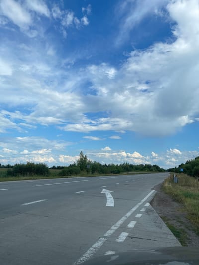This is a ground-level, eye-level, medium shot photograph taken from a vehicle of a two-lane asphalt road in Stremyhorod, Ukraine. The road extends into the distance, curving slightly to the right. White lane markings are visible on the road, including a directional arrow painted on the right lane pointing forward and slightly to the left. The road is bordered on the right by a gravel and grass shoulder, with a blue road sign depicting a straight arrow and a right-turn arrow. Dense green trees and bushes line the left side of the road, and a field of grass is visible behind them. The sky above is a vibrant blue, filled with scattered white cumulus clouds. The scene appears to be a rural or suburban area, likely on a clear day with good visibility. There are no people or animals visible in the image.