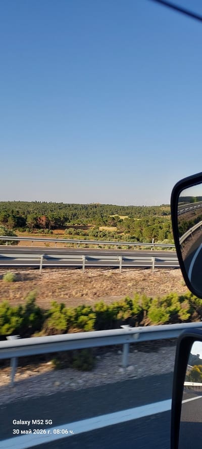 The image depicts a view from a vehicle on a highway. The scene includes a clear blue sky and a highway with a guardrail. On the other side of the guardrail is a landscape with trees and potentially a field. In the distance, there are wind turbines visible. Side mirrors of the vehicle are partially visible in the image. Based on the metadata on the image itself, it's noted that the picture was taken with a Galaxy M52 5G on May 30th, 2026. While Tébar, Spain is mentioned, there isn't specific information in the image to corroborate this location definitively.
