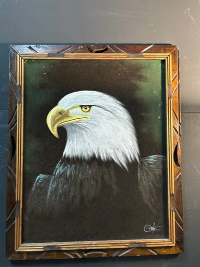 The image displays a framed painting of a bald eagle's head and upper body, rendered in profile, gazing towards the left. The painting appears to be done on a dark, possibly velvet or fabric, background, which is predominantly black with subtle hints of dark green in the upper left and center, suggesting a shadowy environment.

The eagle's head is depicted with bright white feathers, contrasting sharply with the dark background. Its eye is a vivid amber or yellow, encircled by a reddish-brown ring, and has a distinct dark pupil. The beak is prominent, colored in a golden yellow hue with a dark spot near the tip, possibly representing a nostril. The feathers of the eagle's body and wings extend from the head towards the right, transitioning from white into darker shades, likely brown or black, and gradually fading into the dark background, with some lighter strokes hinting at the texture of the flight feathers.

In the upper right corner of the painting, a signature or artist's mark is visible. It appears to be a stylized "Joz" or similar, written in white and partially enclosed within a faint circle.

The painting is housed in a rectangular wooden frame. The frame has a two-tiered design: an inner band of lighter-colored wood with horizontal grooved details, and an outer, darker brown band featuring carved geometric patterns, specifically diagonal lines and V-shapes. The frame shows signs of age and wear, particularly on the left side and bottom edge, where there are noticeable chips and missing pieces of wood, revealing the lighter timber underneath. The damage suggests that the item is vintage or has been handled extensively over time.