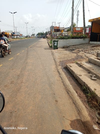 The image shows a street in Ado Ekiti, Nigeria, taken from the perspective of a person riding a motorcycle. The street is paved and has a concrete sidewalk on the right side. There is a green trash can on the sidewalk, and a sign that reads "Crystal Special" in the background. There are several buildings visible on the opposite side of the street, as well as trees and other vegetation. The motorcycle is driving on the left side of the street, and the rider is looking ahead. There are several other vehicles in the distance, traveling in the same direction as the motorcycle. The sky is overcast, but there is no rain. The ground is covered in dirt, and there are several small pieces of debris scattered about. The scene is typical of a street in a developing city. The coordinates provided, 7.649° N, 5.226° E, correspond to a location in Ado Ekiti, Nigeria, close to the intersection of Aramoko Road and the Federal Polytechnic, Ado-Ekiti. This location is identified by the text "Aramko, Nigeria" at the bottom of the image.