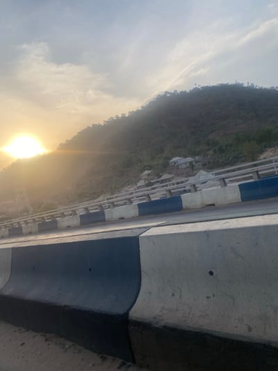 This image depicts a scenic view from a road or bridge winding through a hilly or mountainous landscape, likely during either sunrise or sunset. The overall impression is one of natural beauty combined with infrastructure.
In the foreground, taking up the right side of the frame, is a substantial concrete barrier or wall, which appears to be part of the roadside or bridge structure. This barrier is primarily grey, with a distinct dark blue or black painted section curving along its bottom edge. Higher up on this concrete surface, there are small, dark circular indentations.
Extending horizontally across the mid-ground, a road or bridge is visible, marked by a robust guardrail running alongside it. This guardrail is constructed from concrete blocks, alternately painted white and dark blue or black, creating a repetitive striped pattern that recedes into the distance. Below and to the left of the road, a steep hillside covered in dense, dark green and brown foliage is prominent. Several lighter-colored structures or buildings with slanted roofs are nestled into the side of this hill, appearing somewhat dwarfed by the surrounding nature. Further down the slope, a faint silhouette of a power line tower can be discerned.
The background on the left side of the image is dominated by the sky and a low-lying, intensely bright sun. The sun's strong glare creates a brilliant, warm yellow and orange glow that diffuses across the lower sky and obscures some details of the distant landscape. Above this glow, the sky transitions into a pale blue-grey, scattered with a few delicate, wispy clouds.
The time of day is clearly either early morning or late evening due to the sun's low position and the atmospheric glow it creates. The weather appears clear, though somewhat hazy directly around the sun. There are no people or discernible text visible in the image.