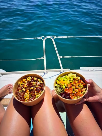 This vertically oriented image captures a serene scene aboard a boat or yacht near Ros Na Rí, Ireland. Two individuals are depicted, primarily by their tanned legs and hands, enjoying poke bowls.

In the upper right, a hand with pink nail polish holds a colorful poke bowl. This bowl contains a vibrant medley of diced salmon, avocado, edamame, bright yellow mango, and green seaweed salad, sprinkled with sesame seeds. Below it, another poke bowl is visible, resting near a second pair of tanned legs. This bowl features diced tuna, avocado, and a dark sauce, also garnished with sesame seeds.

The setting appears to be a sunny day, with clear sunlight casting distinct shadows on the legs, indicating warm and pleasant weather. To the left of the frame, the deep blue-green waters of the sea are visible, showing gentle ripples. Elements of the boat's rigging, including ropes and metal structures, along with the white deck, are present, confirming the maritime location. The overall scene suggests a relaxed and enjoyable meal on the water. There is no visible text in the image.