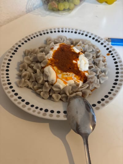 A close-up, eye-level, medium shot captures a plate of manti, a Turkish dish of dumplings, served with yogurt and a spiced oil topping. The manti are small, crescent-shaped, and light gray in color. They are arranged on a white plate with a decorative border of alternating black and gray dots. A dollop of thick white yogurt sits in the center, topped with a vibrant red and orange spiced oil. A shiny metal spoon rests on the white surface of the table in the foreground, slightly to the right of the plate. In the upper right corner of the image, a portion of a blue lighter is visible. The background is out of focus, showing a white countertop and a blurred object that appears to be a bunch of green grapes in a clear plastic bag. The lighting suggests an indoor setting, possibly a kitchen or dining area. The scene appears to be located in Oberasbach, Germany.