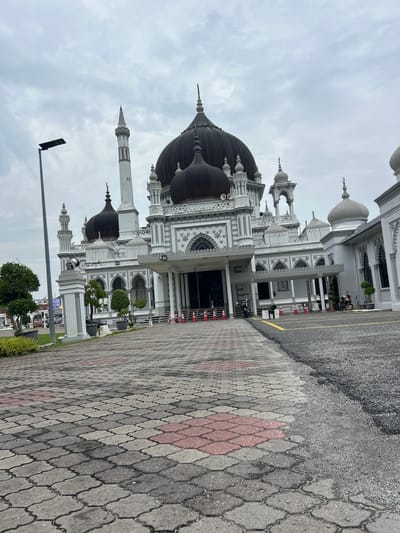 This image shows the exterior of the Zahir Mosque in Alor Setar, Malaysia. The mosque is a prominent landmark, characterized by its white facade, multiple domes, and a tall minaret. The foreground features a patterned paved walkway, with asphalt to the right. Several orange traffic cones are visible along the edge of the walkway, suggesting a temporary barrier or designated area. Two motorcycles are parked near the entrance of the mosque. The sky is overcast, indicating a cloudy day. Trees and some potted plants are also present on the grounds. In the background, the urban environment of Alor Setar can be glimpsed, with buildings and a street visible. The perspective of the image is from a low angle, looking up towards the grand architecture of the mosque.