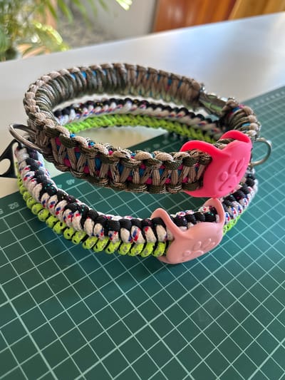 The image shows three handmade paracord dog collars stacked on a green cutting mat. The collars have various color patterns, including camouflage, neon green, black, white, and pink. Two of the collars are adorned with bright pink paw-shaped charms. The background is blurred, showing a plant and a white surface. The image was captured in Coslada, Spain.