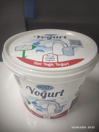 A white plastic tub of yogurt is displayed on a white surface. The tub has blue and red text and graphics. The lid of the tub shows the brand name "DOST Yogurt" in blue lettering. Below this, the text "Tam Yağlı Yoğurt" is written in red lettering on a red stripe. There is also a green sign with white text that reads "Süt Yağı % 4.0 Protein % 4.0". The side of the tub also features "DOST Yogurt" and the weight "3000g". The background is blurred and appears to be an indoor setting. A timestamp in the bottom right corner reads "16/06/2026 20:52". There are no people or animals visible in the image. The location is not specified in the image itself, and therefore cannot be determined.