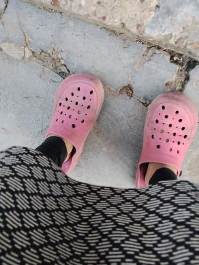 This media file is a close-up, over-the-shoulder shot of a person's feet, wearing pink Crocs and dark leggings, standing on a cracked concrete pathway. The pathway appears to be outdoors, with some dirt and debris visible in the cracks. The lighting suggests it is daytime, possibly late afternoon given the soft shadows. The scene is simple and everyday, focusing on the footwear and the ground. There are no discernable emotions or specific events taking place. The location is Cîrpești, Moldova.