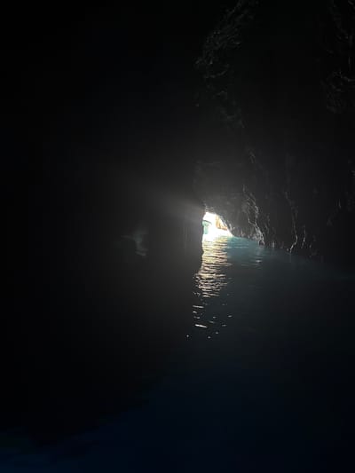 The image depicts a dark cave entrance with bright sunlight illuminating the water inside, located in the city of Komiža, Croatia. The cave walls are rough and textured, with the water appearing to be a deep blue color. Light rays are visible piercing through the darkness, creating a reflective surface on the water. The scene is tranquil and serene, suggesting a natural and untouched environment.
