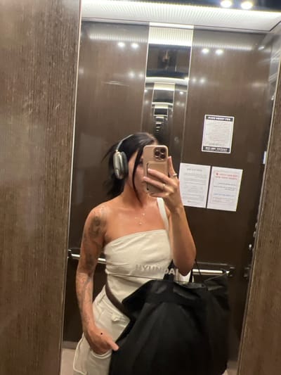 A female subject is depicted taking a mirror selfie inside an elevator located in 강서구, South Korea. She wears a light-colored, one-shoulder top with "HYUNDAI" printed on the front, light-colored bottoms, and silver over-ear headphones. Her left arm, visible to the viewer, is extensively tattooed. She holds a light-colored smartphone to capture the image and carries a large black bag, with a brown strap visible across her torso. The elevator interior consists of mirrored walls, reflecting linear ceiling lighting and a textured wood-grain side panel. Multiple rectangular white notices, displaying Korean text and a '02-' area code phone number, are affixed to the mirrored surface behind her.