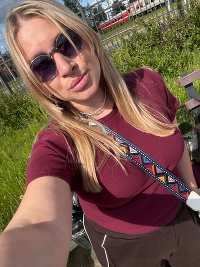 A bright, angled outdoor selfie features a woman with long, sun-kissed blonde hair, fair skin, and plump pink lips, wearing round sunglasses. The text "P-Force P" is visible on the left lens of her sunglasses. She is dressed in a close-fitting, ribbed maroon or burgundy top and dark brown pants or a skirt with white trim details. A prominent, multicolored patterned strap, decorated with geometric shapes in hues of blue, orange, red, and white, crosses her torso. A delicate white beaded necklace is also visible around her neck.
The setting is outdoors, characterized by vibrant green grass and partial sunlight, indicating a sunny day. In the background, behind a decorative black metal fence, a train with red and white carriages is partially visible, suggesting the photo was taken near a railway line or station. A wooden bench is also present in the upper left corner of the frame. The overall scene depicts a casual moment outdoors on a clear day, within the context of Malaya Vishera, Russia.