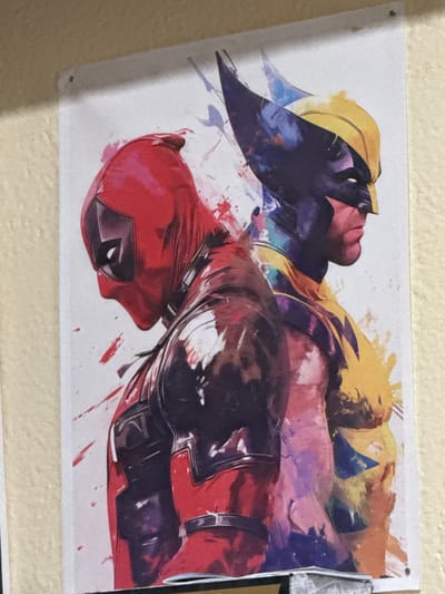 The image depicts a poster on a beige wall. The poster features artwork of Deadpool and Wolverine in a dynamic, possibly combative or collaborative, pose. Both characters are in full costume with Deadpool in his signature red and black and Wolverine in his yellow and blue. The artwork is rendered in a style with brushstrokes and splatters, giving it a painterly feel. There is no indication of the location being Parvomai, Bulgaria.