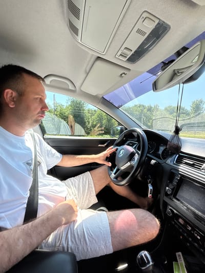 A middle-aged man, wearing a white t-shirt and white shorts, is driving a car. He is looking forward, his hands on the steering wheel. The car's interior is visible, including the dashboard, steering wheel, gear shift, and rearview mirror. The exterior scenery shows trees and a clear blue sky, suggesting a sunny day. The image appears to be taken from inside the car, looking towards the driver. This scene is located in the city of Arefino, Russia.