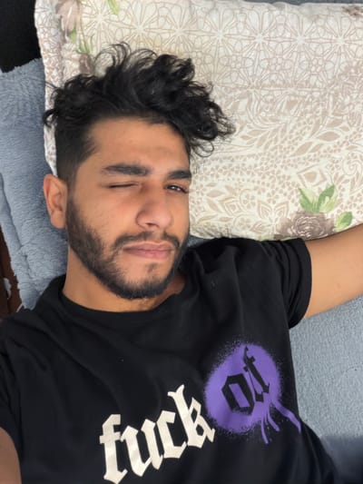 A medium shot shows a man with dark, curly hair and a beard winking at the camera. He is lying on his side, resting his head on a beige and white patterned pillow with a green floral design in the corner. He is wearing a black t-shirt with a large, stylized white word and a purple emblem on the upper left. The man appears to be on a gray blanket or cushion. The location is unknown, with no indication of being in Wirges, Germany.