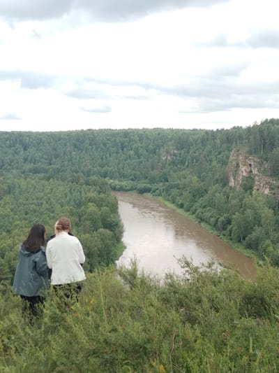 Two individuals, one wearing a dark teal rain jacket and the other a white jacket, stand on a grassy, overgrown hillside overlooking a wide, muddy river that winds through a dense forest. The forest covers rolling hills and cliffs, with a rocky outcrop visible on the right side of the frame. The sky is overcast with heavy, gray clouds. The scene appears to be a natural landscape, possibly a park or a viewpoint in the vicinity of Idrisovo, Russia, given the description.