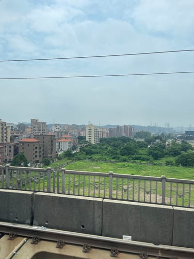The image, taken from a train, shows a view of Dongguan, China. In the foreground, there are train tracks and a concrete barrier. Beyond the barrier is a decorative metal fence and a field of green grass. In the middle ground, there are numerous apartment buildings and some lush green trees. The background is filled with more apartment buildings, power lines, and a hazy sky. The overall scene suggests an urban or suburban environment with a mix of residential buildings and green spaces.