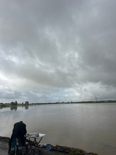 The image depicts an outdoor scene in Oumazza, Morocco, under a heavily overcast sky dominated by dense grey clouds. A single individual, observed from behind, is bent forward, facing a small white rectangular folding table positioned on a muddy bank at the edge of a large, calm body of water. The person wears a dark, long overcoat and blue trousers with prominent reflective yellow-green stripes near the ankles, along with dark boots. On the table, a silver teapot, at least two clear glass cups, and a dark rectangular tray are visible. The immediate ground around the table is muddy and dark, with scattered rocks and what appears to be a crumpled blue plastic bag. The expansive body of water is a dull grey-brown, exhibiting minimal ripples and reflecting the overcast sky and distant landscape. Along the distant left shoreline, green fields and a cluster of trees are visible, with their reflections mirrored in the water. Stretching across the horizon on the right side of the image is a line of electricity pylons, with indistinct structural elements visible further in the distance.