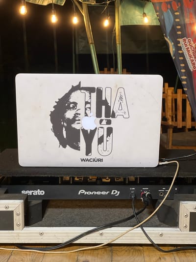 The image presents a nighttime scene in Mururiini, Kenya, centered around a DJ setup. A silver MacBook laptop is prominently displayed, its lid adorned with a large black and white sticker. The sticker features a stylized face and the text "THAU" (with the Apple logo cleverly replacing the 'A') and "WACIURI." The laptop is connected via cables to a professional Pioneer DJ mixer, which also bears the "serato" branding. The mixer itself is housed within a sturdy flight case, indicating portable and robust equipment for events.
In the background, illuminated string lights cast a warm, ambient glow against the darkness, confirming the nighttime setting. These lights are strung across what appears to be scaffolding or a wooden frame, suggesting an outdoor or temporary event venue. Partially visible at the top left is a colorful banner with the text "GuChoki Rui MUKARO - A Kikuyu Culture Podcast." This banner provides a strong cultural context, indicating a podcast or event celebrating Kikuyu culture, consistent with the location in Kenya. The overall arrangement suggests that a musical performance or cultural event is either in progress or about to begin, creating an atmosphere of entertainment and community engagement.